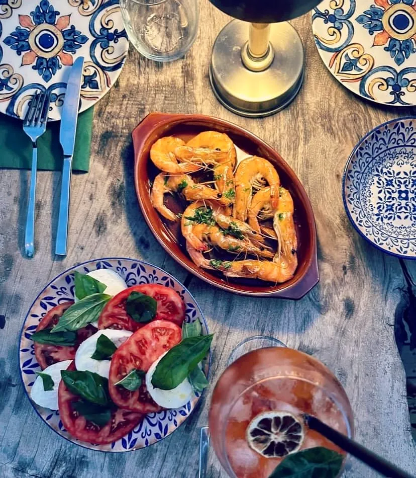 Trattoria Il Mandarino'da ahşap bir masada, Caprese salatası, fesleğen ve mozzarella ile dilimlenmiş domatesler, yeşilliklerle süslenmiş oval bir tabakta pişmiş karidesler ve desenli seramik tabaklarla kurulmuş bir sofra düzeni.