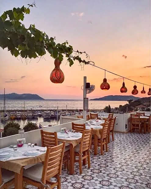 Gün batımının romantik tonlarına bürünen gökyüzünün altında, masmavi deniz ve marinadaki teknelerin eşlik ettiği bir teras restoranında, üzüm asmalarından sarkan oyma kabak lambaların aydınlattığı zarif masalar ve ahşap sandalyelerle keyifli bir akşam yemeği atmosferi.
