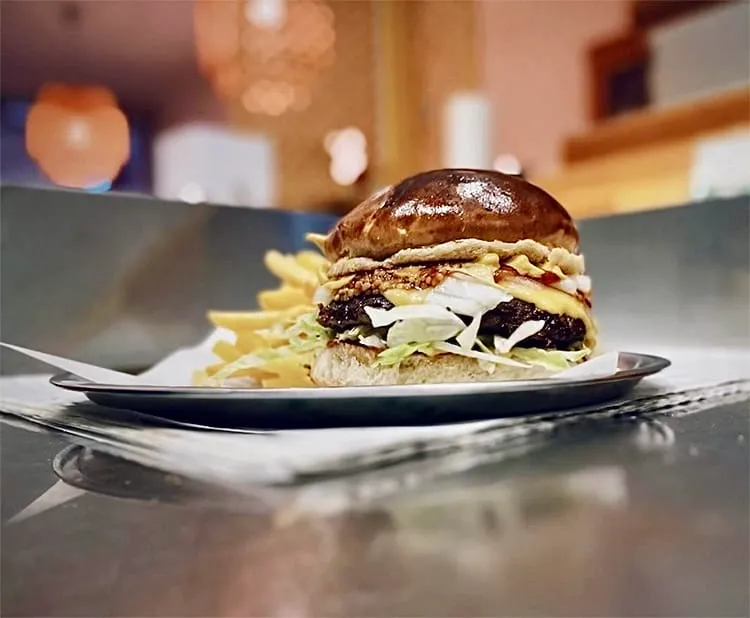 Parlak, hafif kızarmış brioche ekmeği arasında kalın köftesi, erimiş peyniri ve bol maruluyla sunulan, yanında çıtır çıtır patates kızartması olan iştah açıcı bir burger, loş ışıklı bir ortamda metal bir tabakta duruyor.
