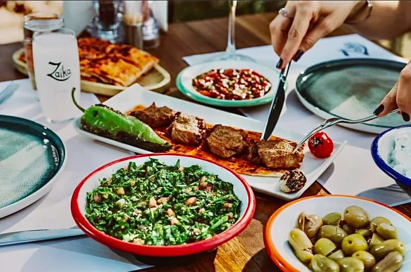 Şiş kebaptan et kesen bir kişinin ellerinin göründüğü bir masada, Zaika logolu rakı bardağı, bol yeşillikli salata, zeytin, acılı ezme ve cacık gibi zengin Türk mutfağı mezeleri ile unutulmaz bir akşam yemeği ziyafeti sunuluyor.