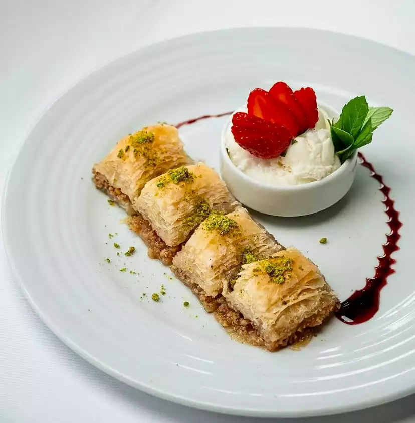 Antalya'nın zengin mutfağını yansıtan beyaz bir tabakta sunulan, üzeri bol Antep fıstığı ile süslenmiş dört dilim şerbetli baklava, yanında çilek dilimleri ve nane yapraklarıyla süslenmiş bir kase vanilyalı dondurma ile iştah açıcı bir gastronomi şöleni sunuyor.