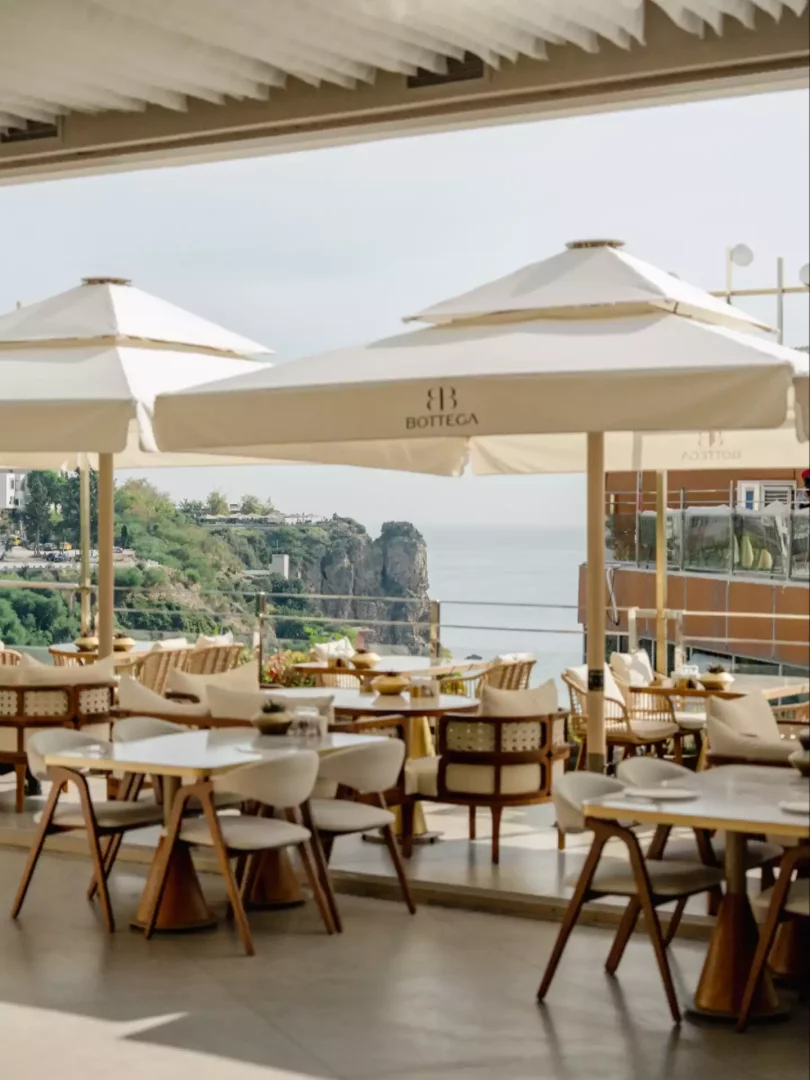 Masmavi denize ve etkileyici kayalıklara bakan, Bottega logolu bej şemsiyelerin gölgelediği ferah bir açık hava restoran terası, modern ahşap detaylı beyaz masa ve sandalyelerle misafirlerini ağırlamaya hazır, huzurlu bir atmosfer sunuyor.