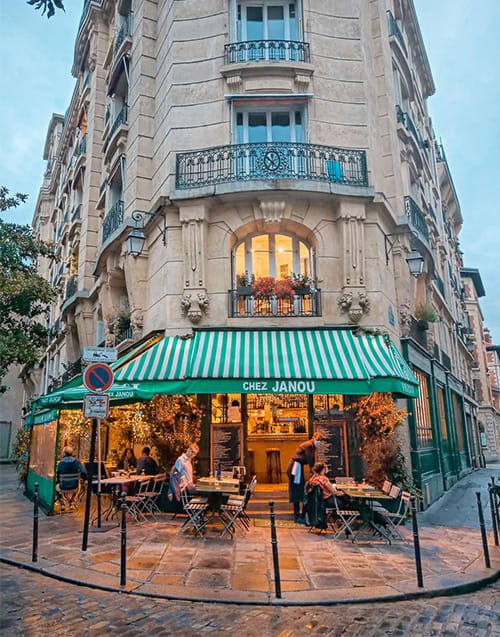 Paris'in bilinen yerel lezzet duraklarından Chez Janou'nun, yeşil beyaz çizgili tente altındaki kaldırımda oturan misafirleriyle, sıcak ışıklarla aydınlanan iç mekanından ve üst katlardaki süslü balkonlarından yansıyan samimi atmosferi, şehrin en iyi restoranları arasında otantik bir deneyim sunuyor.