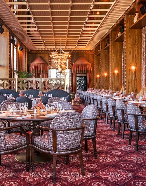 Zengin desenli kırmızı halılar, ahşap panelli duvarlar ve aynalı tavan ile oryantalist bir atmosfer sunan Dar Mima, özenle hazırlanmış masaları ve işlemeli sandalyeleriyle Paris restoranları arasında Mağrip esintileri taşıyan şık bir yemek salonunu gösteriyor.