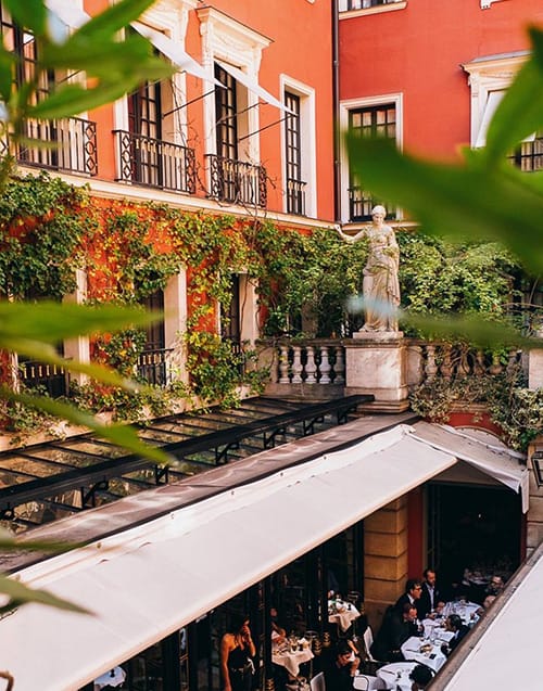 En İyi Paris Restoranları başlığındaki Hotel Costes'in büyüleyici atmosferini yansıtan, kırmızı binasının yemyeşil sarmaşıklarla bezeli cephesi önünde, zarif bir heykelle süslenmiş beyaz tenteli açık hava restoranında keyifli yemek anları yaşayan konuklar.