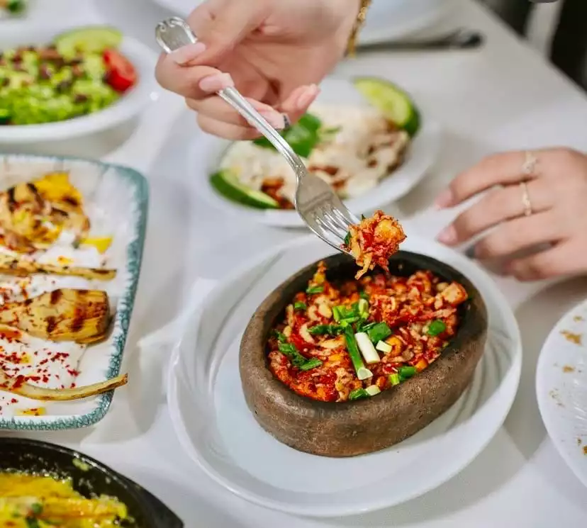 Bir restoran masasında, bir elin çatalıyla güveçte servis edilen, üzerinde taze yeşil soğanlar bulunan kızarmış kalamar tavanın bir porsiyonunu alırken, arka planda ızgara enginar kalpleri ve salata gibi Akdeniz mutfağının diğer zengin lezzetleri Antalya'nın önde gelen restoranlarından birindeki seçkin menüyü gözler önüne seriyor.