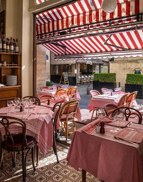 Bir Paris restoranının içinden görünen manzara, kırmızı-beyaz ekose masa örtülü samimi masaları ve hasır sandalyeleriyle başlarken, dışarıdaki sokağa doğru uzanan ikonik çizgili tentelerin altındaki hareketli kaldırımı ve diğer klasik Paris restoranlarını göstererek şehrin En İyi Paris Restoranları atmosferini yansıtıyor.