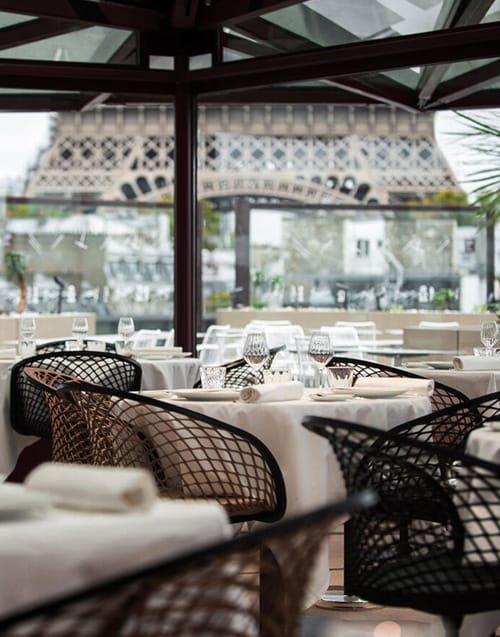 Geniş cam duvarları sayesinde Eyfel Kulesi'nin büyüleyici manzarasına nazır, beyaz örtülü şık masaların ve modern kafes tasarımlı sandalyelerin özenle yerleştirildiği bu Paris restoranında, misafirleri enfes bir gastronomi deneyimi bekliyor.