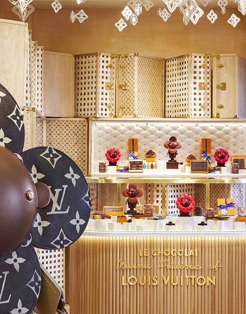 Louis Vuitton Cafe'nin Maxime Frédéric imzalı lüks tezgahında, markanın ikonik monogram desenleriyle süslenmiş büyük bir top figürünün yanında, zarif sunumlarla sergilenen çiçek ve geometrik şekilli çikolatalar ve tatlılar, Paris restoranları içinde özgün bir gastronomi deneyimi sunuyor.