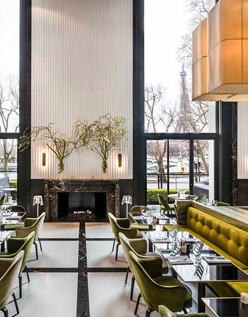 Eyfel Kulesi'nin uzaktan göründüğü pencere önünde, modern ve şık bir atmosfere sahip Monsieur Bleu Paris restoranının iç mekanı; yüksek tavanlar, yeşil kadife sandalyeler ve koyu renk mermer detaylarla dolu yemek masalarını gözler önüne seriyor.