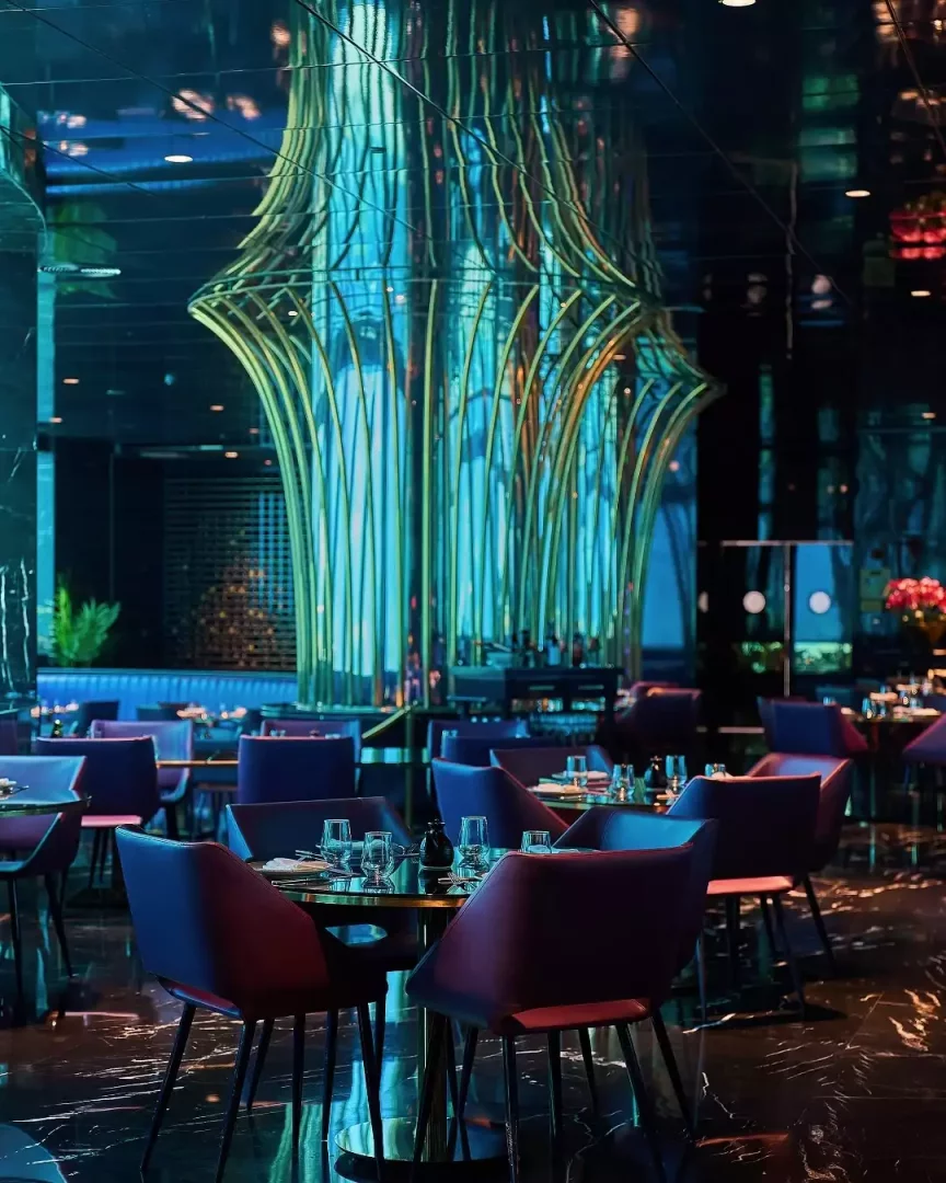 Modern bir restoranın büyüleyici iç mekanı, mavi ve turkuazın hakim olduğu aydınlatmasıyla adeta bir su altı dünyası hissi uyandırıyor; merkezde yükselen, altın renkli ve neon mavi ışıklarla vurgulanmış, deniz yaşamını anımsatan sanatsal armatür, mor koltuklu şık masaların üzerinde yansımasıyla mekana eşsiz bir derinlik katıyor.