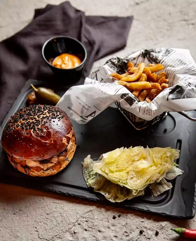 Antalya'nın en beğenilen hamburger restoranlarından birinde şık bir sunumla servis edilen, siyah susamlı hamburger ekmeği, çıtır patates kızartması, özel sosu ve yanında taze göbek marul ile turşusuyla iştah açıcı bir hamburger tabağı.