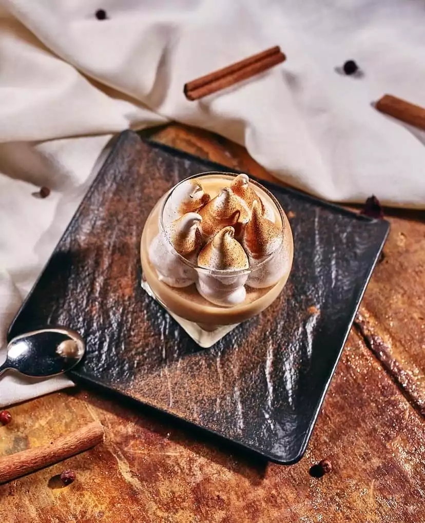 Antalya'da keyifli bir gastronomi yolculuğunu tamamlamak üzere, üzerinde hafifçe kızartılmış beze tepeleri bulunan açık kahverengi bir kremalı tatlının, koyu renkli dokulu bir tabakta şık sunumu, tarçın çubukları ve baharatlarla çevrili.
