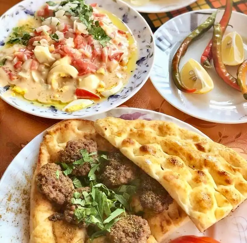 Yakın çekimde bir masada, domates, soğan ve haşlanmış yumurta dilimleriyle zenginleştirilmiş tahinli Antalya usulü piyaz, taze yeşilliklerle pidelerin üzerinde sunulan baharatlı şiş köfteler ve yanında acı biber ile limon dilimlerinin bulunduğu tabaklar, Antalya mutfağının özgün lezzetlerini gözler önüne seriyor.