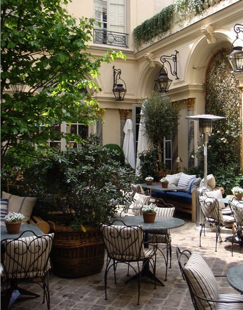 Paris'in en iyi restoranlarından birinin avlusunda, ferforje sandalyelerin çizgili minderlerle süslendiği, masalarda küçük çiçek saksılarının bulunduğu, yemyeşil bitkilerle çevrili şık bir açık hava yemek alanı görülüyor.