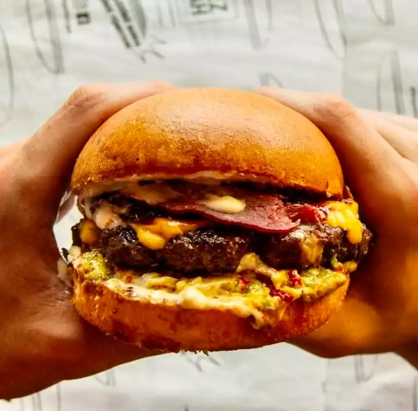 Antalya'nın en iddialı hamburger mekânlarından Tezgah Burger'e ait, çift köfteli, erimiş peynirli, dana pastırmalı ve özel soslarla zenginleştirilmiş, iki elle tutulan lezzetli bir gurme hamburgerin yakın plan fotoğrafı.