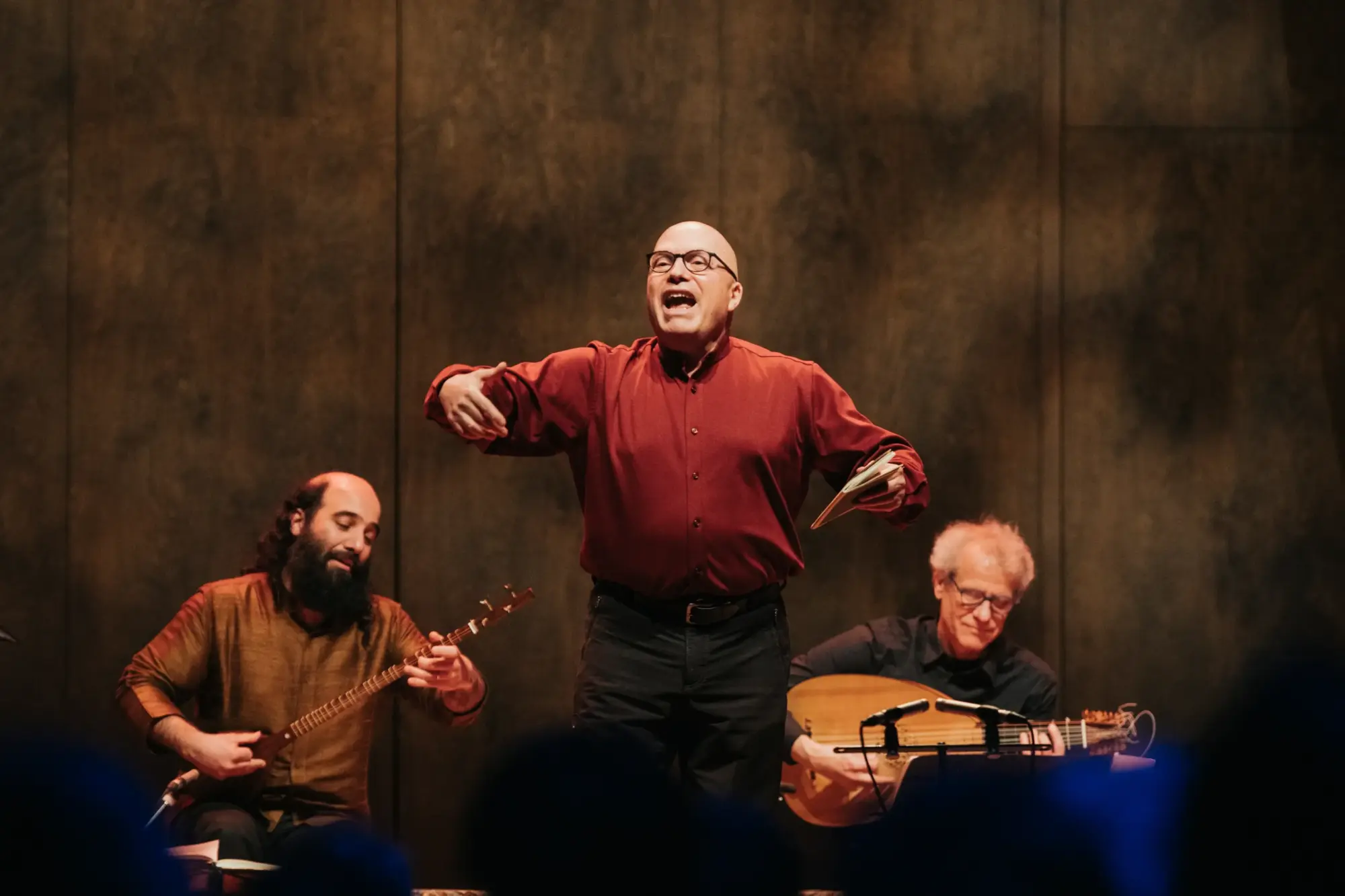Sahne ışıkları altında kırmızı gömlekli bir anlatıcının performans sergilediği, yanında telli çalgılar çalan iki müzisyenin yer aldığı canlı müzik gösterisi.