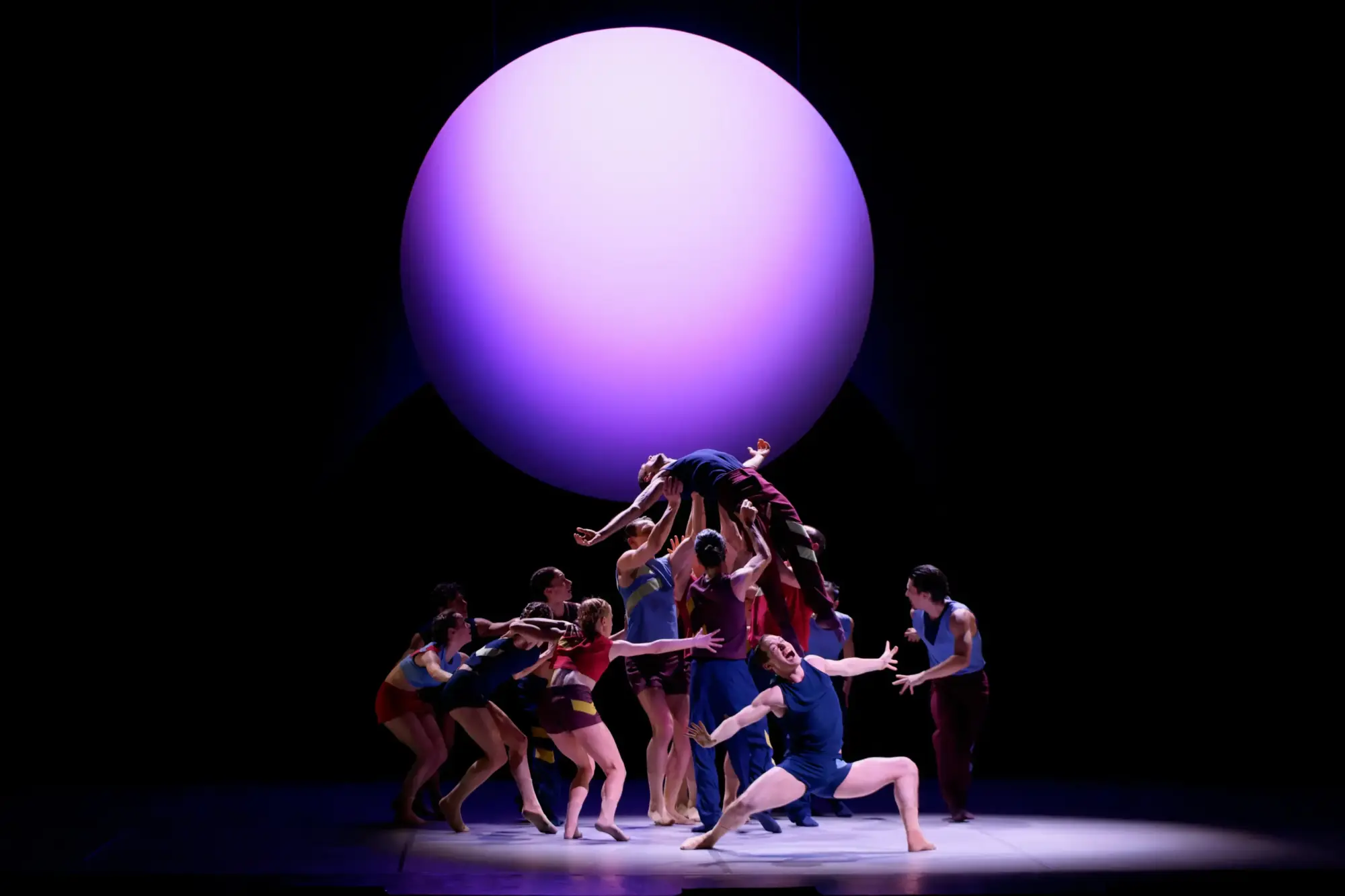 Aterballetto dans topluluğunun sahne performansı; mor ışıkla aydınlatılmış dev yuvarlak dekor önünde grup koreografisi.