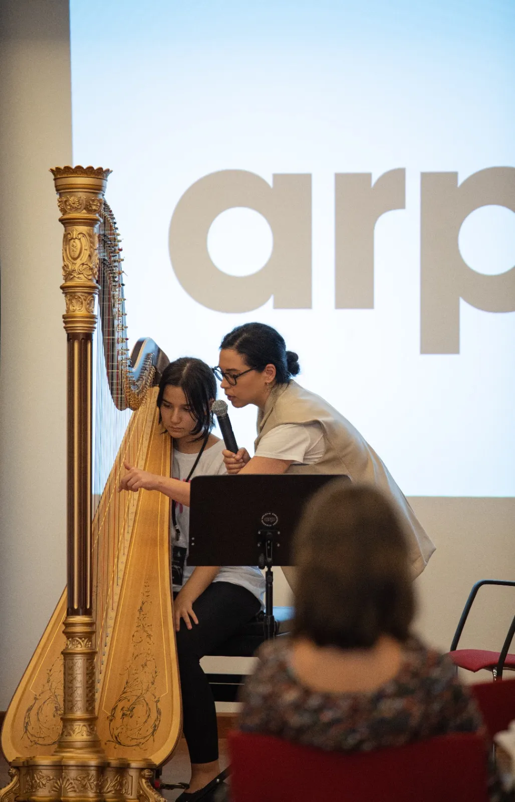Sahne üzerinde altın renkli bir arp başında oturan genç müzisyene mikrofonla rehberlik eden eğitmen; arka planda projeksiyon perdesi ve izleyiciler görülüyor.