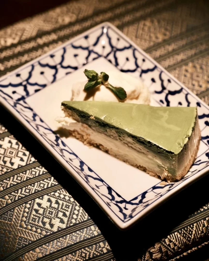 Geleneksel Thai motifleriyle süslü bir tabakta sunulan, üst katmanı matcha veya pandan gibi yeşil renkli, alt katmanı kremalı peynirli ve bisküvi tabanlı cheesecake diliminin yanında nane yaprağıyla süslenmiş bir top krema veya dondurma görülüyor; bu özel Thai tatlısı koyu renkli ve altın işlemeli bir masa örtüsü üzerinde göz alıcı duruyor.