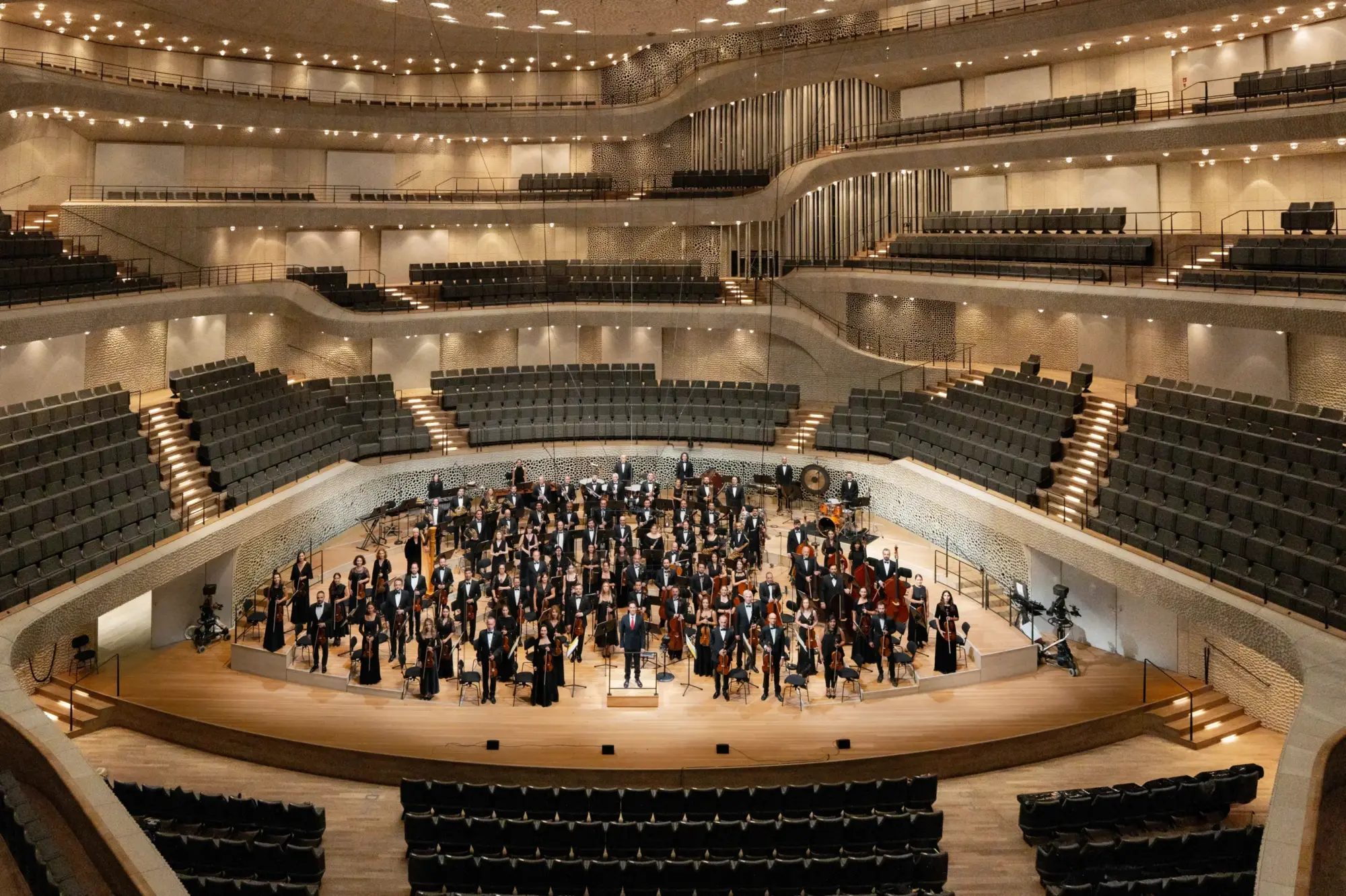 Modern bir konser salonunda sahnede toplanmış büyük bir senfoni orkestrası; çok katlı seyirci balkonları ve geniş ahşap sahne alanı görülüyor.