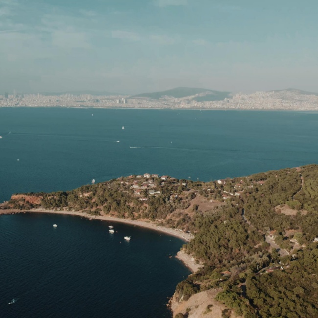 Burgazada'nın yeşil tepeleri, ormanlık alanları ve sahildeki sakin koyunda demirli tekneleriyle birlikte, karşı kıyıda yükselen İstanbul siluetinin panoramik bir görünümü, bu gezi rehberi için adanın huzurlu atmosferini yansıtmaktadır.