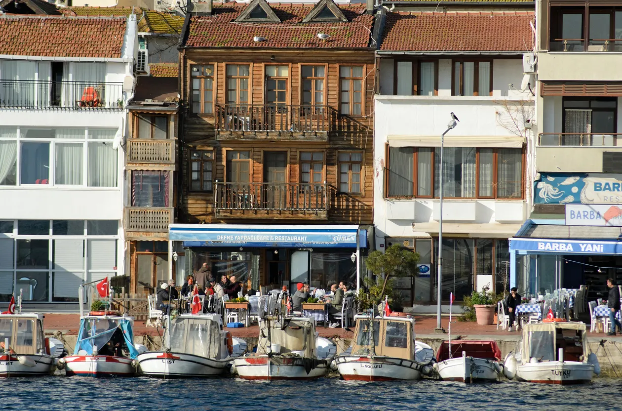 Burgazada'da deniz kenarında sıralanmış geleneksel ve modern mimariye sahip binalar, önlerinde bağlı duran tekneler ve dışarıda yemek yiyen insanlarla dolu restoranlar adanın huzurlu ve canlı atmosferini yansıtıyor.