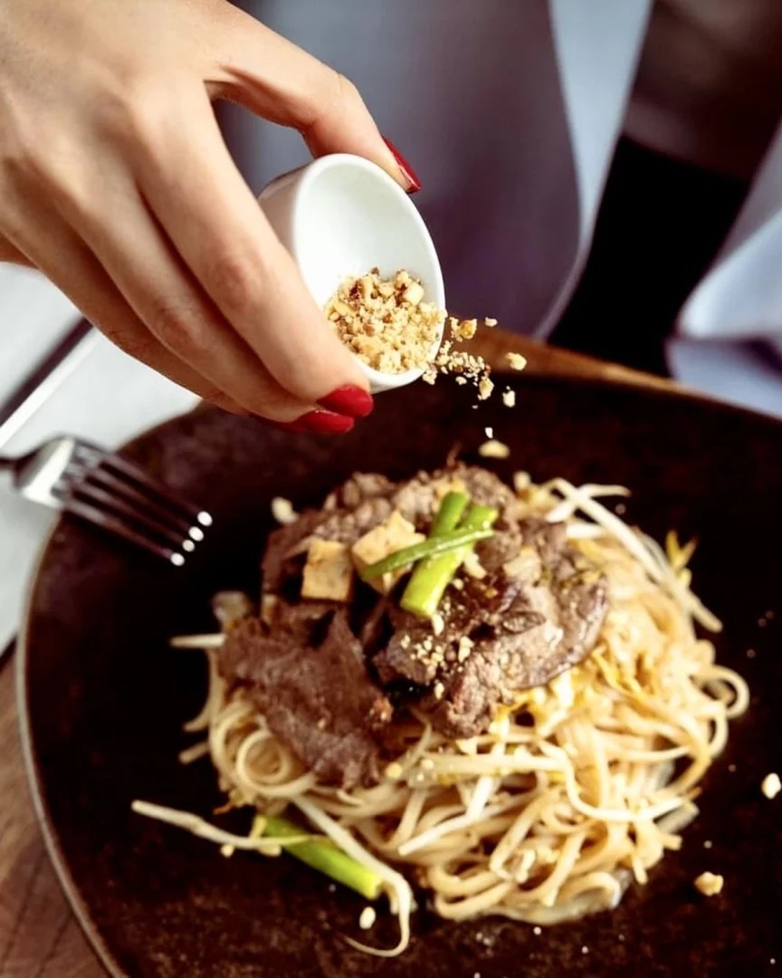 Kırmızı ojeli bir el, küçük beyaz bir kaptan doğranmış yer fıstıklarını, içinde noodle, dana eti dilimleri, soya filizi, yeşil soğan ve tofu küpleri bulunan koyu renkli bir tabaktaki zengin Tayland yemeği üzerine özenle serpiştiriyor. İstanbul’daki Tay restoranları arasında yer alan Çok Çok Thai mekânından bir görünüm.