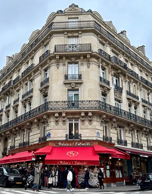 Paris'in klasik mimarisini yansıtan süslü demir balkonlu, bej taşlı bir köşedeki bina, kırmızı tenteleriyle dikkat çeken ve önünde sıra bekleyen insanlarla şehrin en sevilen Paris restoranlarından Le Relais de l'Entrecôte'u gösteriyor.