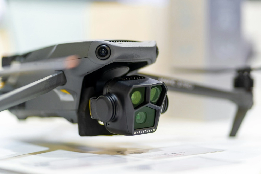Yakın çekimde görünen koyu gri renkli DJI Mavic 4 drone'un ön kısmında, üç belirgin yeşil lensi ve üzerinde HASSELBLAD markası bulunan gelişmiş kamera sistemi net bir şekilde odaklanmış, üst tarafında ise ek bir sensör yer alıyor.