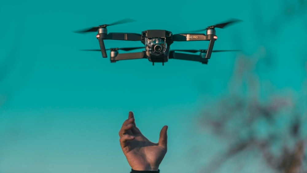 Parlak turkuaz gökyüzünde havada süzülen, hareket halindeki pervaneleri bulanık görünen bir DJI Mavic Pro katlanabilir drone, altında uzanan bir elin üzerinde uçarken çekilmiş detaylı bir görüntü.