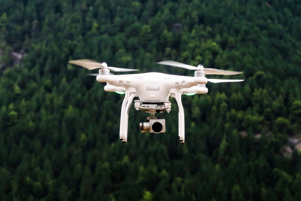 Beyaz renkli DJI Phantom 4 4K drone, pervaneleri döner vaziyette havada süzülürken altında yüksek çözünürlüklü çekimler için tasarlanmış bir kamera taşıyor, geniş yeşil ormanlık alan ise bulanık bir fonda belirginleşiyor.