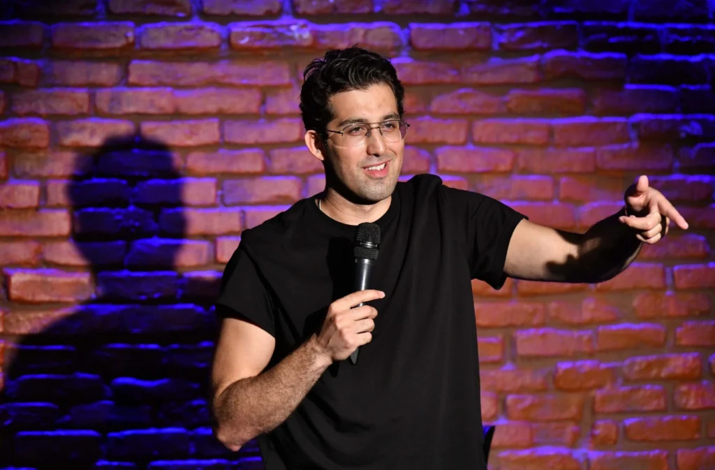 Komedyen Doğu Demirkol, İstanbul'daki bir stand-up etkinliği kapsamında sahnede siyah tişörtü ve gözlükleriyle mikrofonu tutarak tuğla duvar önünde sağa doğru neşeyle işaret ediyor, izleyicisiyle samimi bir etkileşim içinde.