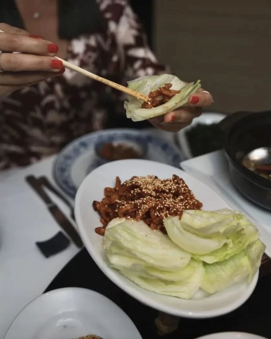 Kırmızı ojeli bir el, yemek çubukları ile marul yaprağına susamlı et yemeğini özenle yerleştirerek bir marul sarması hazırlıyor; beyaz oval tabakta ise bolca aynı yemek ve taze marul yaprakları görülüyor, bu lezzetli Asya mutfağı deneyiminin bir restoranda keyifle yaşandığını gösteriyor. İstanbul Uzak Doğu restoranları arasında yer alan Dragon Restaurant mekânından bir görünüm.