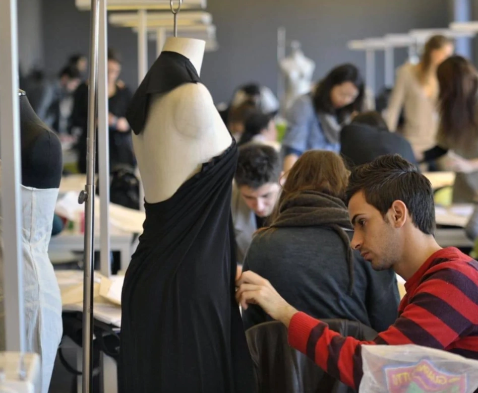 Ön planda askıda siyah dökümlü bir elbisenin prova edildiği cansız mankenlerin arasında, arka planda masa başında ve etrafında dikkatle çalışan çok sayıda öğrencinin yoğun bir moda tasarımı atölyesindeki yaratıcı süreci yansıttığı canlı bir moda okulu ortamı.