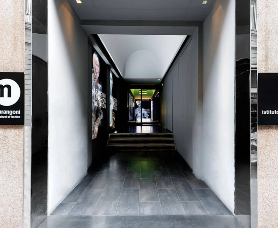 Istituto Marangoni'nin girişini temsil eden modern bir koridor, sol duvarda Marangoni School of Fashion tabelası ve MARANGONI FASHION MUTATION yazan büyük moda posterleri ile dünyanın en iyi moda okullarından birinin dinamik ruhunu yansıtıyor.