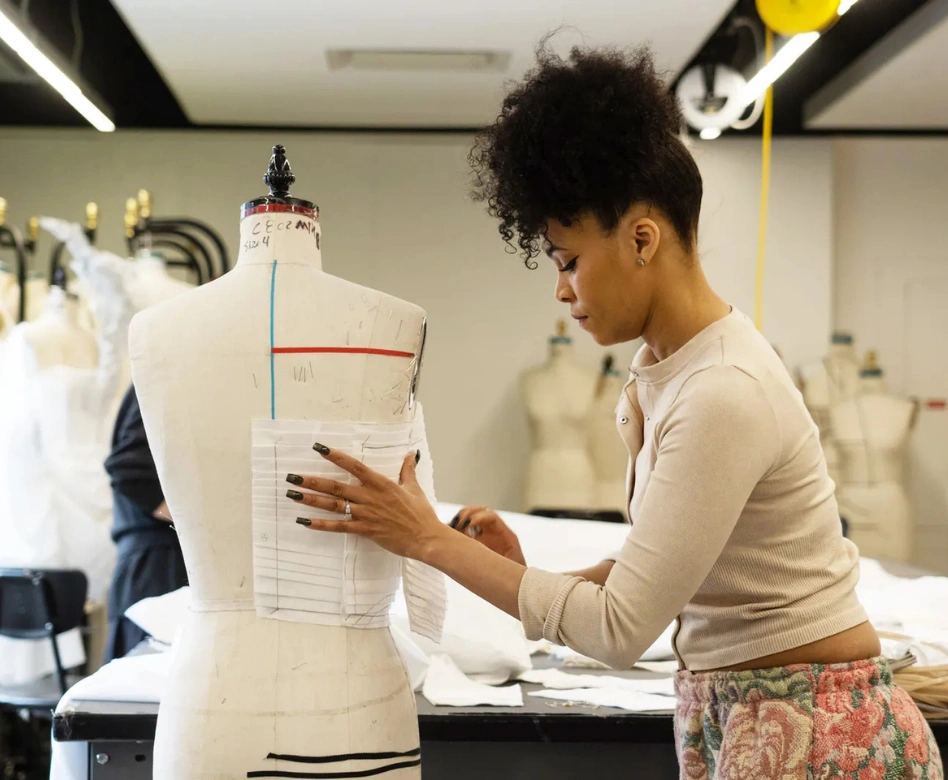 Kıvırcık saçlı genç bir kadın, bir moda atölyesinde, ölçü işaretli bir terzi mankeni üzerinde beyaz bir kumaş parçasını titizlikle çalışarak şekillendiriyor, arka planda ise diğer mankenler ve çalışma masaları bulanık bir şekilde seçiliyor.