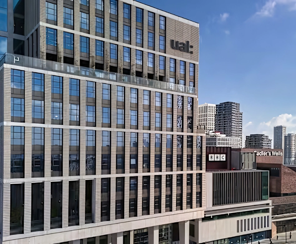 Yüksek, cam pencereleri ve modern gri cephesiyle dikkat çeken, üzerinde belirgin UAL logosu bulunan bu şehir binası, dünyanın en iyi moda okullarından biri olan London College of Fashion'ın bir parçası olarak, arka planda BBC binası ve diğer Londra yapılarıyla şehir manzarasını tamamlıyor.