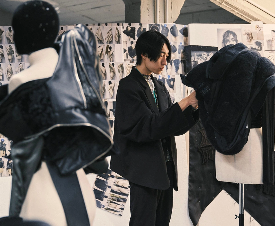 Dünyanın en iyi moda okullarından Polimoda'da bir moda öğrencisi, tasarım stüdyosunda, koyu renkli, dokulu bir kıyafeti cansız manken üzerinde özenle şekillendirirken, arka planda ilham panoları ve taslaklar ile yaratıcı bir çalışma ortamı sergileniyor.