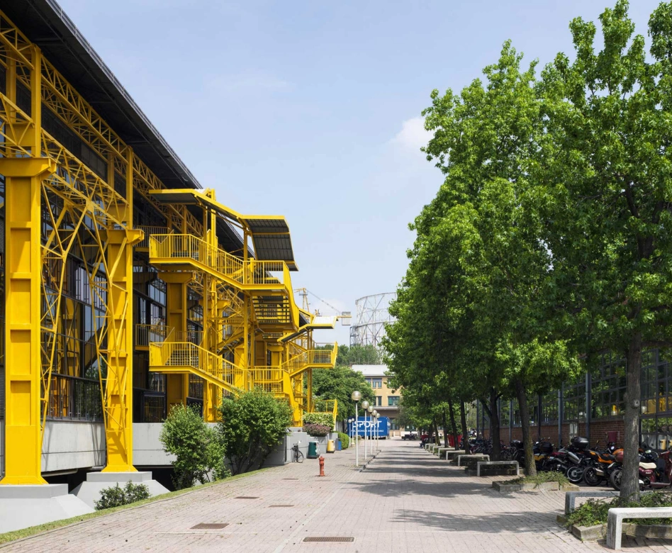 Parlak sarı taşıyıcı elemanları ve dışarıdan erişilen modern merdivenleriyle dikkat çeken, cam cepheli bir eğitim binası, önünde sıralı ağaçlar, taş döşeli bir yürüyüş yolu ve park edilmiş motosikletlerin bulunduğu geniş bir kampüs alanında konumlanmıştır; bu görüntü, dünyanın en iyi moda okullarından biri olarak bilinen Politeknik'in yenilikçi mimarisini ve canlı atmosferini gözler önüne seriyor.