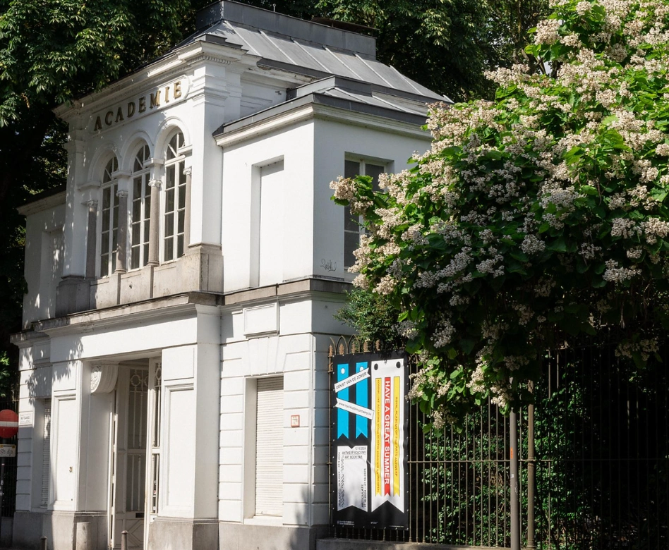 Beyaz cepheli, ACADEMIE yazılı klasik mimarili bir binanın önünde, sağda çiçek açmış beyaz bir ağacın dalları ile kısmen kaplanmış ve üzerinde Antwerp Güzel Sanatlar Akademisi'ne ait bir posterin asılı olduğu demir çitlerin görüldüğü bu detaylı fotoğraf, dünyanın en iyi moda okullarından biri olan Antwerp Güzel Sanatlar Akademisi'nin girişini güneşli bir günde gözler önüne seriyor.