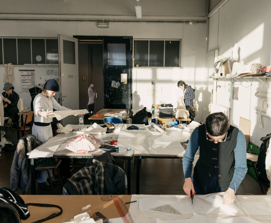Güneşli bir moda atölyesinde, bir öğrenci desen keserken, diğerleri masalarda kumaş ve taslaklar üzerinde yoğun bir şekilde çalışıyor, en iyi moda okullarının tipik yaratıcı ve hareketli atmosferini yansıtıyor.