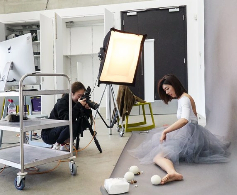 Royal College of Art gibi dünyaca ünlü moda okullarındaki yaratıcı çalışmaları temsil eden bu görselde, gri bir stüdyo fonu önünde oturan bir modelin, sırtı açık beyaz bluz ve hacimli gri tül etekle poz verdiği, bir kadın fotoğrafçının da tripod üzerindeki kamerası ve profesyonel ışık ekipmanlarıyla çekim yaptığı yoğun bir moda çekimi anı yer alıyor.
