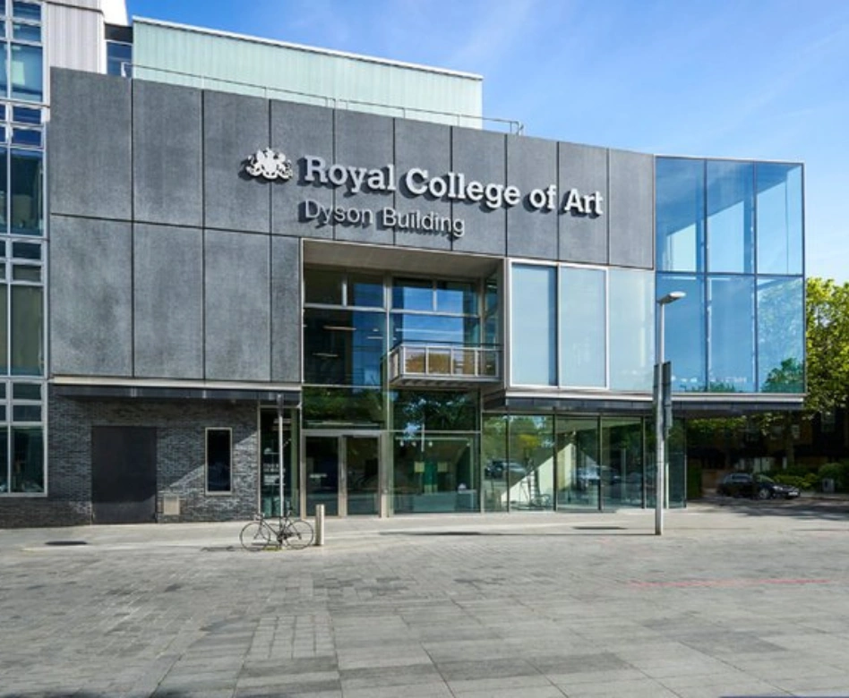 Koyu gri paneller ve geniş cam cepheleriyle dikkat çeken, modern mimariye sahip Royal College of Art Dyson Binası, dünyanın en iyi moda okullarından biri olarak önünde duran bir bisikletle birlikte güneşli bir günde dışarıdan görülüyor.