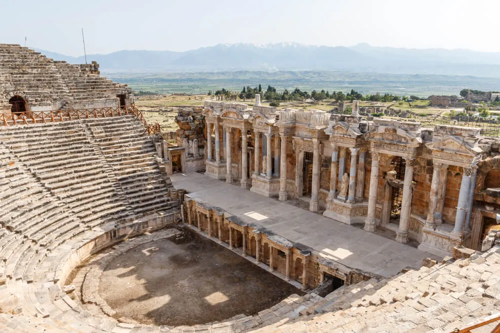 Yüksek bir açıdan çekilmiş bu fotoğrafta, Hierapolis Antik Kenti'nin basamaklı oturma sıralarına ve özenle süslenmiş, sütunlu sahne binasına sahip heybetli Büyük Tiyatrosu, Pamukkale'nin doğa harikasıyla bütünleşen zengin tarihi dokusunu gözler önüne seriyor, arkasında uzak dağlarla çevrili geniş bir ova uzanıyor.