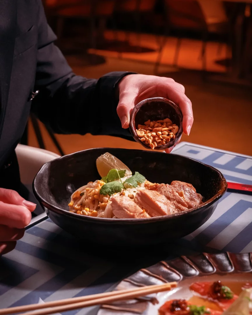 Koyu renk bir kasede sunulan, limon dilimi ve taze kişniş ile zenginleştirilmiş tavuklu pad thai yemeğinin üzerine, Jie Istanbul'daki bir restoranda koyu renk ceketli bir el tarafından küçük bir kaseden özenle ufalanmış fıstıklar serpiştirilirken, masadaki desenli örtü ve yanındaki suşi tabağı şık bir yemek deneyimini gözler önüne seriyor.