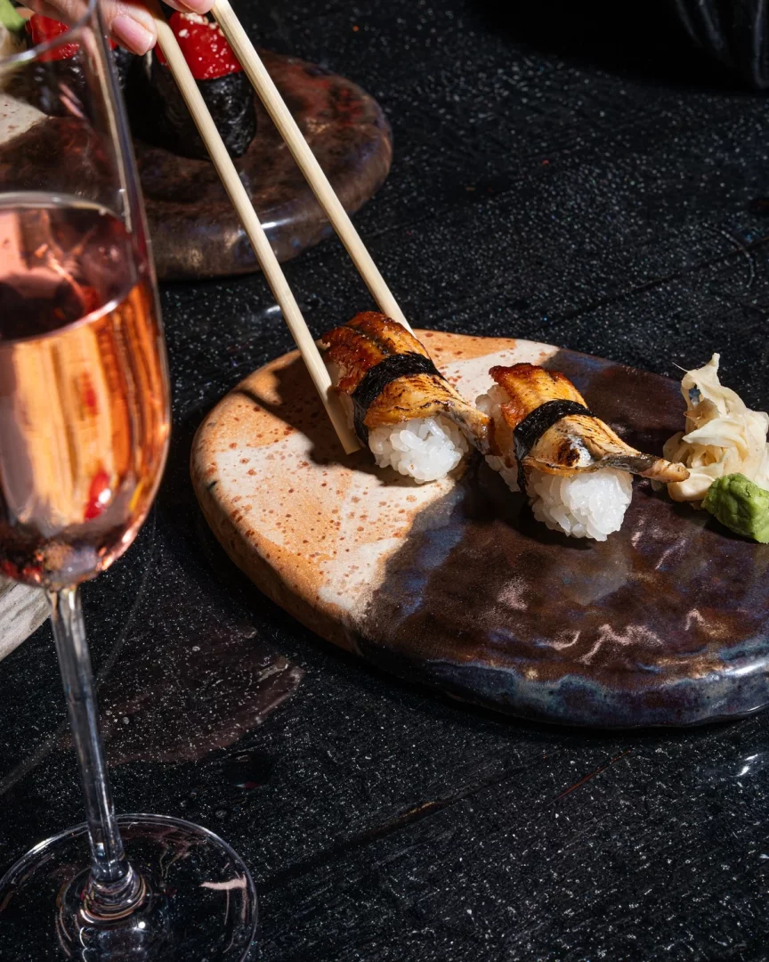 Bir elin yemek çubuklarıyla zarifçe tuttuğu, özel tasarım seramik bir tabağa özenle yerleştirilmiş iki parça iştah açıcı yılan balığı nigiri sushinin yakından çekilmiş görüntüsü; yanındaki kadehte rose şarap, taze zencefil ve wasabi ile tamamlanarak şık bir İstanbul restoran deneyimini çağrıştırıyor.