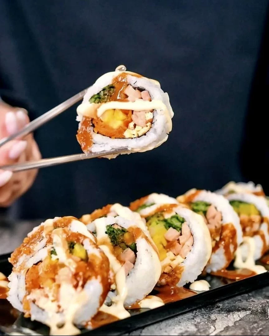 Siyah, uzun bir tabakta duran ve bolca beyaz mayonez ile kahverengi tatlı acı sos gezdirilmiş altı adet Kore Köyü sushi rulosunun görüldüğü bu yakın çekim görselde, bir çift metal çubukla havaya kaldırılan bir dilimin içindeki yeşillikler, havuç, jambon ve omlet gibi çeşitli dolgular dikkat çekiyor. İstanbul’daki Kore restoranları arasında yer alan Koreköy mekânından bir görünüm.