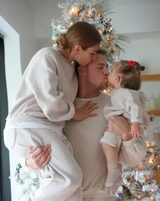 Beyaz ve krem tonlarında uyumlu kıyafetler giymiş bir baba, anne ve bebeğin olduğu bir aile portresi, aile üyeleri birbirlerini öpüyor.