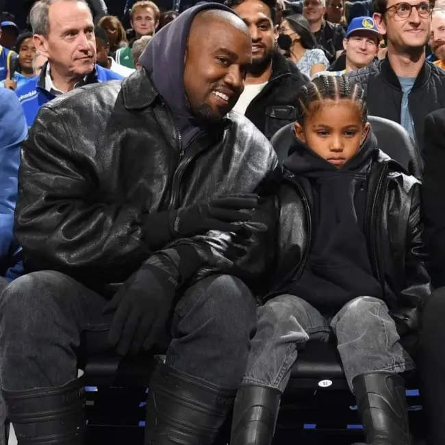 Kanye West ve çocuğunun, babalar ve çocuklar için Mini Me trendine uygun, uyumlu koyu renk giysiler ve botlarla bir spor etkinliğini izledikleri bir an.